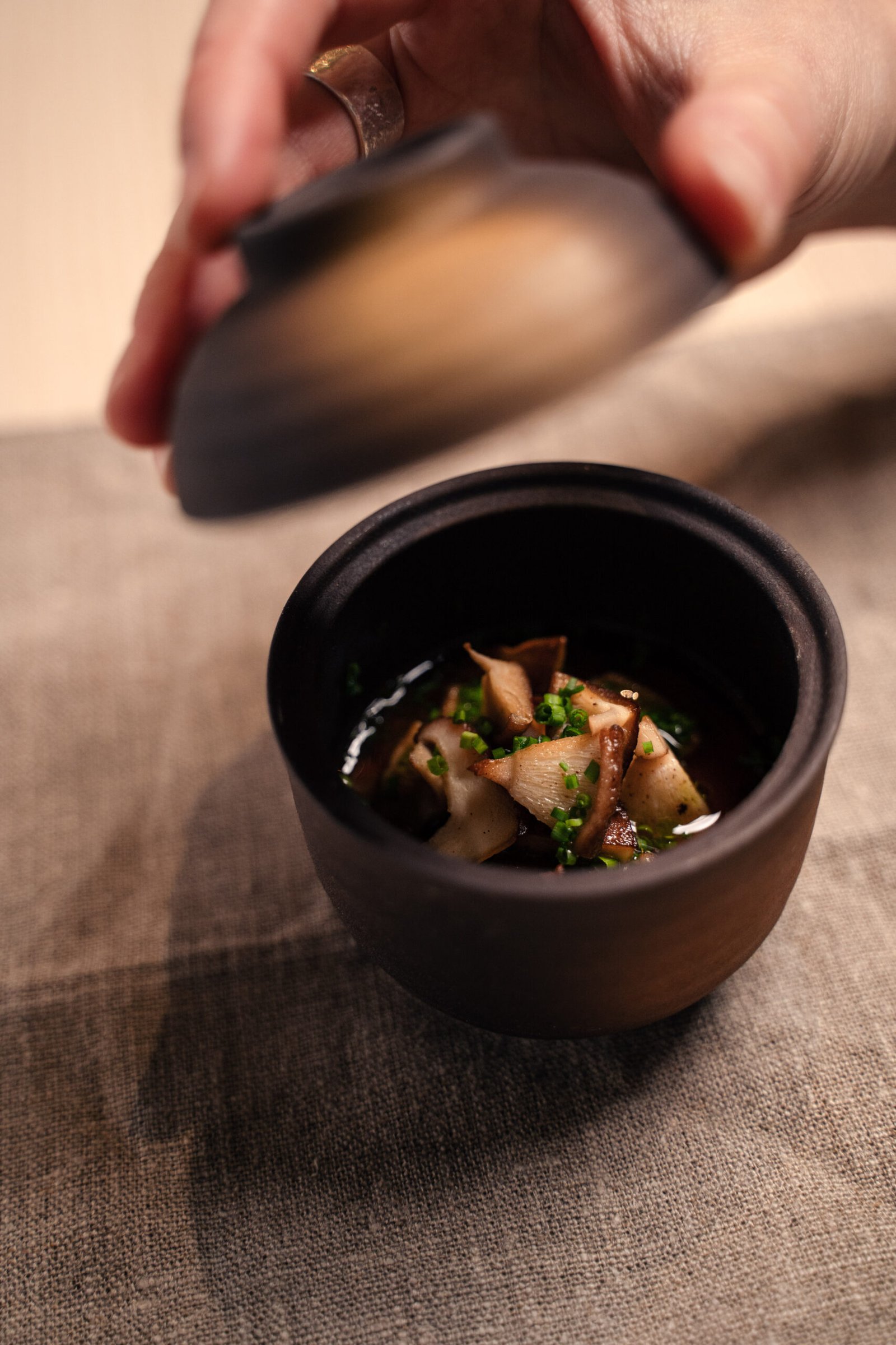 A dark bowl of mushroom broth with chives, lid being lifted